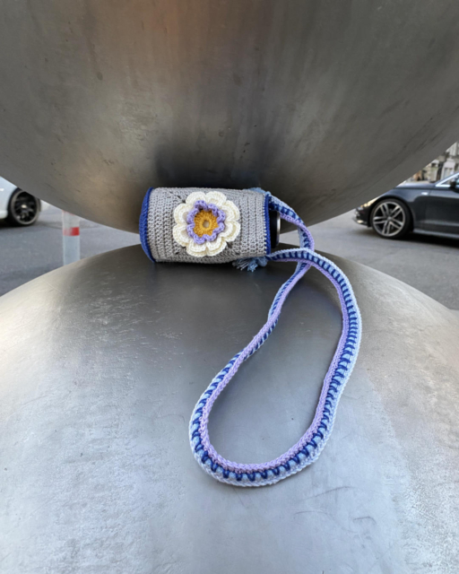 small crocheted bottle bag with flowers lying between two huge metal spheres
