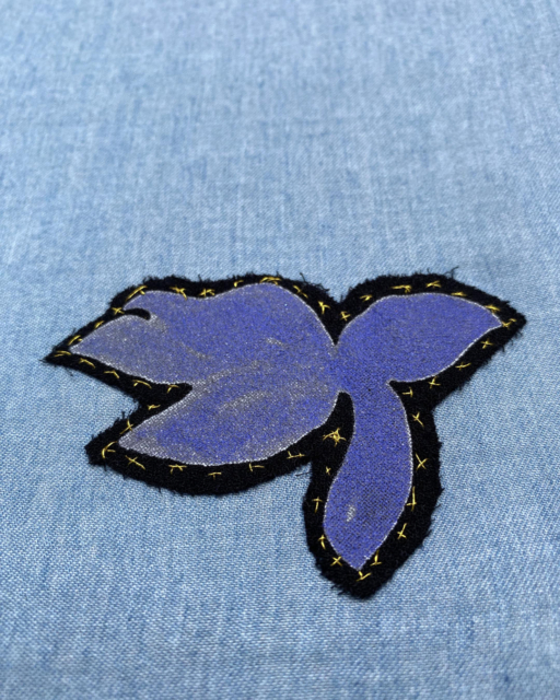 cut out lilac flower stitched on light blue cloth with golden thread